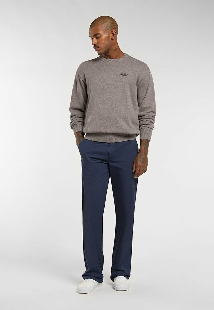 Man with tattoos wearing gray sweater, navy pants, and white sneakers, standing with hands in pockets looking down.