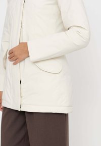 Giacca impermeabile color crema con pattina frontale testurizzata, tasche laterali e design liscio e aderente. Abbinata a pantaloni marroni.