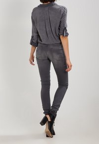Camisa gris de manga larga con puños enrollados, combinada con jeans gris ajustados y botas negras hasta el tobillo, vista desde atrás.