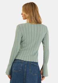 Hellgrüner, gestrickter Pullover mit vertikalen Rippen, gewelltem Saum und ausgestellten Ärmeln, kombiniert mit einem Jeansrock mit hinteren Taschen.