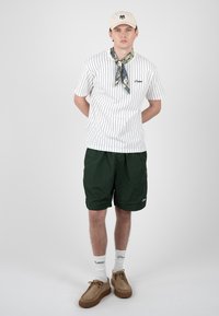 Jeune homme portant un T-shirt blanc à fines rayures, un short vert foncé, une casquette beige, un foulard à motifs autour du cou, des chaussettes blanches et des chaussures marron, debout devant un fond gris.