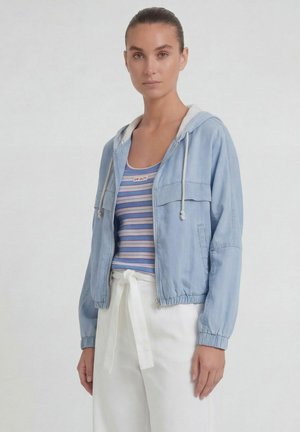 Femme portant un sweat à capuche zippé bleu clair sur un débardeur rayé bleu et un pantalon blanc taille haute avec ceinture nouée, debout devant un fond uni.