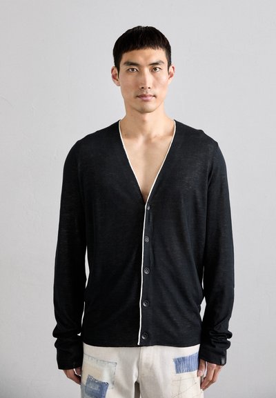 Cardigan noir à manches longues avec un col en V, bord blanc le long de la patte de boutonnage et fermeture à boutons. Porté sur un pantalon clair avec des patchs.