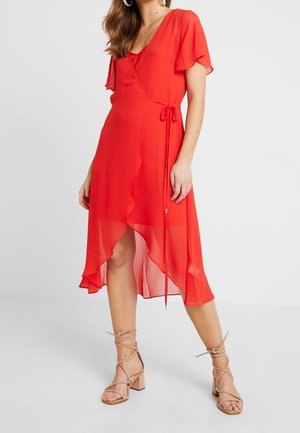 Žena v červených zavinovacích šatech s volánovými rukávy a asymetrickým lemem, doplněných béžovými sandály na blokovém podpatku se šněrováním.