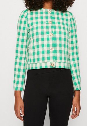 Femme portant une veste à carreaux verte et blanche boutonnée avec des boutons dorés et un pantalon noir, debout, les bras détendus le long du corps.