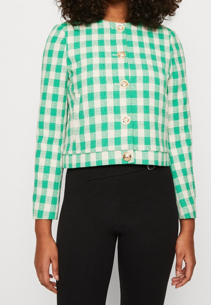 Femme portant une veste à carreaux verte et blanche boutonnée avec des boutons dorés et un pantalon noir, debout, les bras détendus le long du corps.