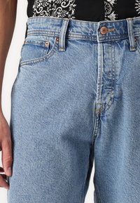 Jeans din denim de un bleu deschis, cu multiple buzunare, închidere cu nasture și cusături rezistente. Țesătura are un finisaj ușor texturat.