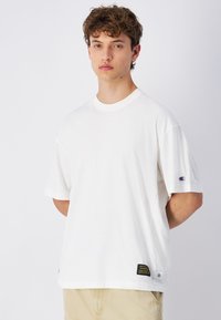 T-shirt bianco a maniche corte in cotone, con collo rotondo, logo sulla manica sinistra e un'etichetta con testo nero e giallo all'orlo.