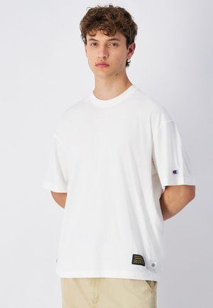 T-shirt bianco a maniche corte in cotone, con collo rotondo, logo sulla manica sinistra e un'etichetta con testo nero e giallo all'orlo.