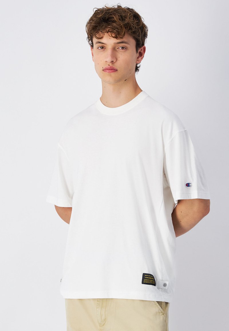 T-shirt bianco a maniche corte in cotone, con collo rotondo, logo sulla manica sinistra e un'etichetta con testo nero e giallo all'orlo.