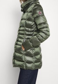 Green puffer jacket with glossy finish, quilted design, high collar, and side pockets. Features a circular logo on the left sleeve.