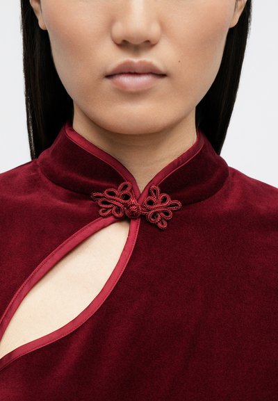 Top din catifea bordeaux cu guler mandarin, având o decupare laterală și închideri intricate cu noduri brodate. Textură netedă și culoare bogată.