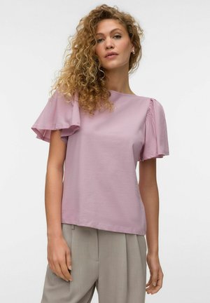 Kvinde med krøllet hår iført en lysegrå lilla top med flagrende korte ærmer og beige bukser med høj talje mod en ensfarvet baggrund.