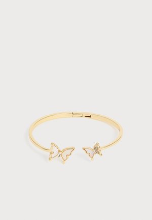 Brazalete abierto dorado con dos extremos en forma de mariposa, uno adornado con pequeños cristales transparentes, sobre un fondo liso y claro.