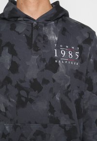 Grau camoufliertes Hoodie mit Klappkragen und zwei Knöpfen. Zeigt das Grafikdesign "TOMMY 1985 HILFIGER" in Weiß und Rot. Aus weichem Baumwollmaterial.