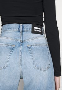 Person som bär ljusblå denimjeans med en svart lapplabel och en svart långärmad topp, visad bakifrån från midjan och neråt.