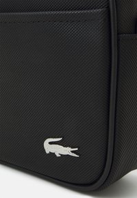 Svart texturerad väska med en silverfärgad krokodillogo, släta kanter och en strukturerad design. Materialet verkar hållbart och stiligt.