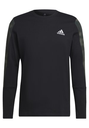 Personne portant un maillot Adidas noir à manches longues avec des bandes vert olive sur les manches, debout les mains jointes.