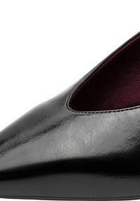 Gros plan sur la pointe pointue d'une chaussure en cuir noir brillant avec des coutures visibles et une doublure intérieure en tissu rouge foncé.