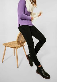 Pantaloni neri slim-fit, maglione viola a maniche lunghe a coste e una sciarpa bianca, abbinati a stivali neri alla caviglia, seduto su una sedia di legno intrecciato.