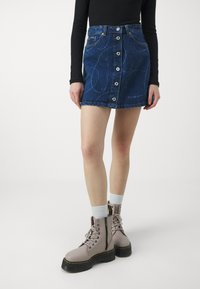 Denimkjol i mörkblått med knapprad fram, fem fickor och diskreta sömnadsmönster. Kombinerad med ljusrosa kängor och vita strumpor.