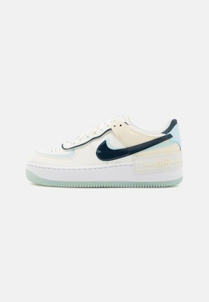 Sneaker in pelle bianca con accenti blu navy, suola verde menta e punta forata. Presenta un'etichetta "AIR" ben visibile sul tallone.