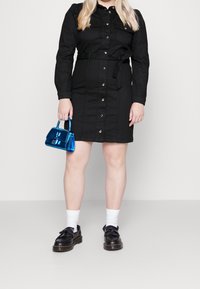 Robe chemise en denim noir avec une taille ceinturée, des boutons à l'avant et deux poches poitrine. Tient un petit sac à main bleu brillant ; chaussures noires et chaussettes blanches.