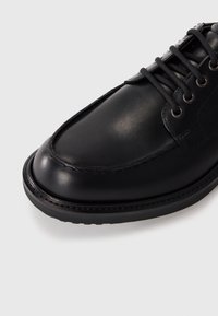 Schwarzer Lederschuh mit abgerundetem Zehenbereich, genähten Akzenten und Schnürdesign. Verfügt über eine strukturierte Oberfläche und eine schwarze Gummisohle.