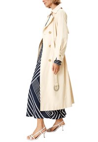 Gabardina beige con herrajes dorados, de longitud hasta la rodilla, con un diseño clásico. Se lleva sobre un vestido de rayas azul marino y blancas, combinado con tacones blancos de tiras.