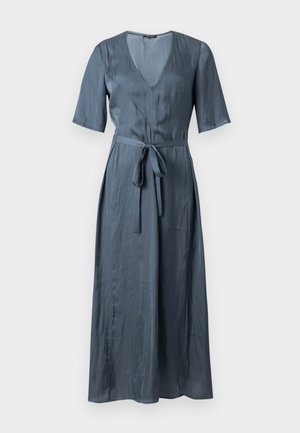 Žena v modrých saténových midi šatách s krátkymi rukávmi, výstrihom do V, opaskom v páse a čiernymi koženými čižmami do polovice lýtka stojaca na jednofarebnom pozadí.