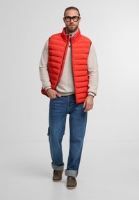 Rote wattierte Weste mit hohem Kragen, kombiniert mit einem hellgrauen Pullover und blauen Jeans. Die Weste hat einen Reißverschluss vorne und horizontale Nähte.