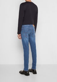 Modré denimové džíny s úzkým střihem, s zadními kapsami a kontrastním šitím. Nosič má na sobě černou košili s dlouhým rukávem a černé boty.