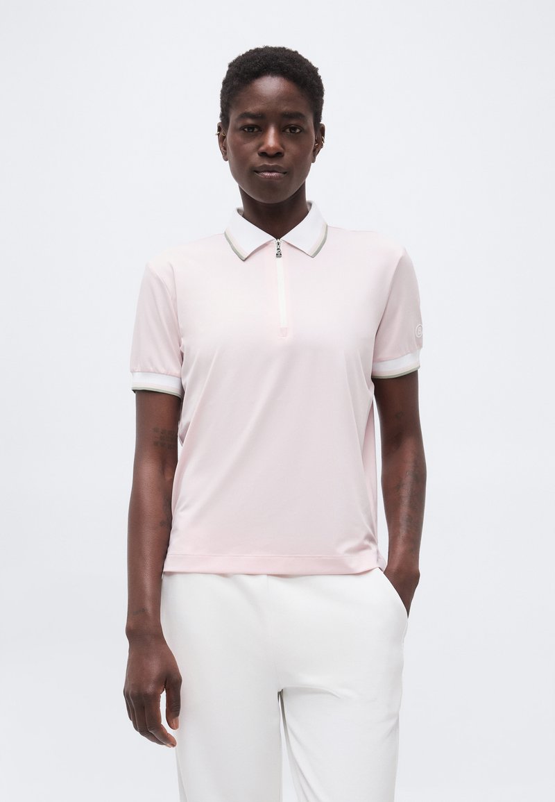 Kvinde med kort krøllet hår iført en lyserød kortærmet polo-shirt og hvide bukser, stående med den ene hånd i lommen.