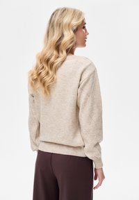 Beżowy, dziergany sweter o luźnym kroju, z diamentowym wzorem na rękawach i prążkowanym obszyciem. Połączony z ciemnobrązowymi spodniami.