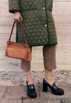Persoon draagt een groene gewatteerde jas, geruite cropped broek, zwarte loafers met blokhak, en houdt een bruine leren en suède handtas naast zich vast.