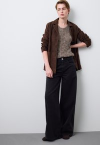 Brauner Wildleder-Blazer über einem strukturierten Strickpullover und weiten schwarzen Jeans. Das Outfit zeichnet sich durch eine lockere Passform, neutrale Farbtöne und subtile Details aus.