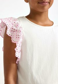 Hvid bomulds skjorte med rund halsudskæring, der har rosa blonde flæser ved ærmerne med hulmønstre og broderede detaljer.