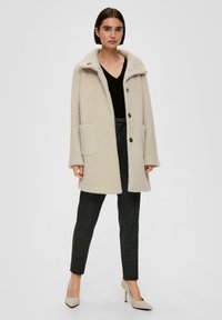 Beige wollen jas met een gestructureerde afwerking, brede kraag en knoopsluiting. Bevat twee zijzakken en is gecombineerd met donkere broeken en hakken.