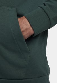 Dark green sweatshirt featuring a textured fabric, ribbed cuffs, and a front pocket. Close-up of a hand in the pocket.
