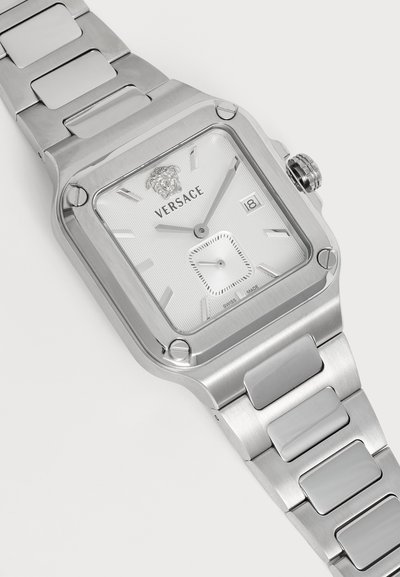 Montre-bracelet Versace argentée avec cadran carré, affichage de la date, logo et détail tête de lion sur un bracelet à maillons métalliques.