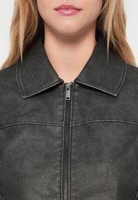 Veste en simili cuir noire avec un col structuré et fermeture éclair, présentant une finition texturée et des coutures sur la poitrine.