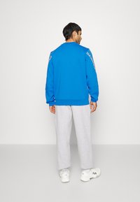 Sinine dressipluus pikkade varrukatega, soonikkoes randmetes ja ümar kaelus; koos heledate hallide dressipükste ja valgete tossudega.