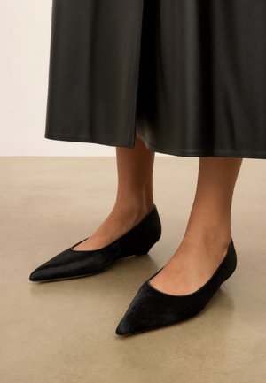 Vrouw die zwarte laaggehakte schoenen met een puntige neus en een zwarte leren midi-rok draagt, staand op een beige vloer.