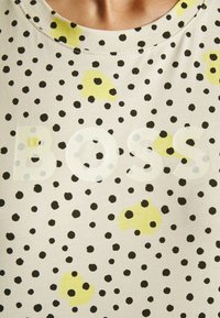 Close-up af en beige skjorte med sorte polkaprikker, gule abstrakte pletter og ordet "BOSS" trykt i store gennemsigtige bogstaver hen over brystet.