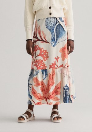 Jupe mi-longue avec des imprimés de corail et d'algues marines bleues et de coquillages, portée avec un pull crème et des sandales blanches ouvertes sur une personne à la peau foncée.