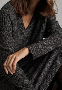 Ciemnoszary sweter dziany z głębokim dekoltem w serek, szerokimi rękawami i miękką fakturą, narzucony na nogi z wyraźnie widoczną dłonią spoczywającą na nich.