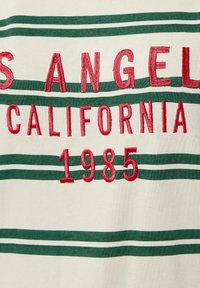 Cremefarbene Stoff mit grünen horizontalen Streifen und rotem gesticktem Text "LOS ANGELES CALIFORNIA 1985." Weiche Textur, lässiges Design.