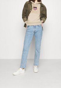 Felpa beige con logo, giacca in velluto a coste verde oliva, jeans azzurri chiari e sneakers bianche. Outfit casual a strati con colori contrastanti.