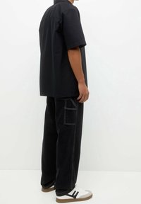 Hombre con una camisa negra de manga corta holgada, pantalones anchos negros con costuras blancas y zapatillas blancas con rayas negras.