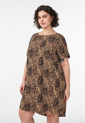 Moteris su garbanotais plaukais vilki trumpomis rankovėmis leopardinės spalvos suknelę, stovėdama priešais baltą foną, rodydama tatuiruotę kairėje rankoje.